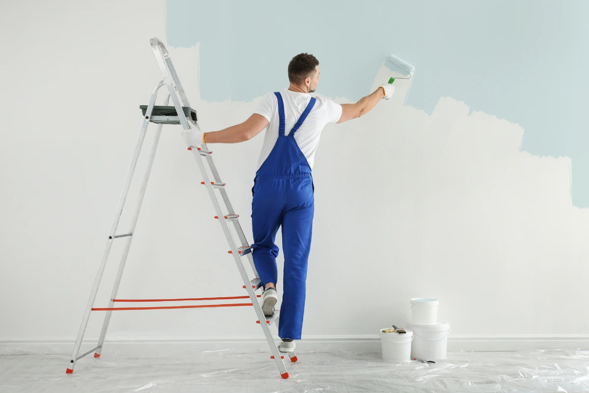 Painter in blue overalls painting a wall white.